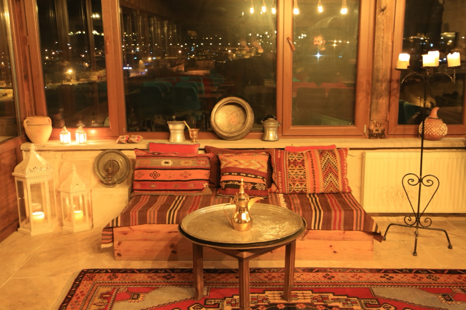 Gece ışıkları görünen geniş camlı oturma salonu; kilim yastıklar, bakır sini masa ve çaydanlık